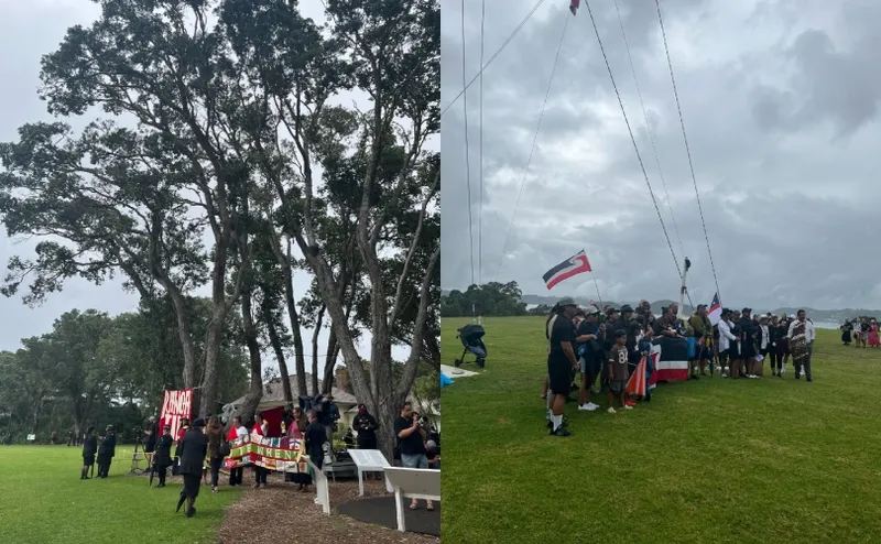 A group of people gather outdoors near tall trees and on open grass, holding banners and flags, including the Tino Rangatiratanga flag, during a cloudy day event or protest.