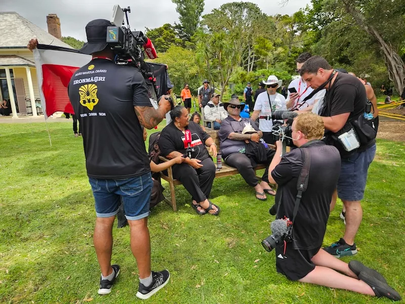 A group of people, some with cameras and filming equipment, gather around two people sitting on chairs outdoors on grass. Trees and a white building are in the background. One seated person holds a red flag.