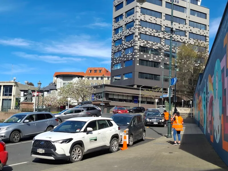 A busy city street with parked and moving cars, a tall modern building in the background, and two people wearing orange safety vests standing on the sidewalk near a colorful mural.