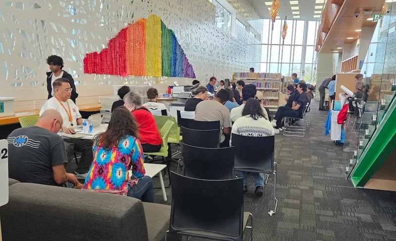 A group of people sit at tables and work on laptops in a modern, open library space with colorful wall art and large windows, while others browse books or walk nearby.