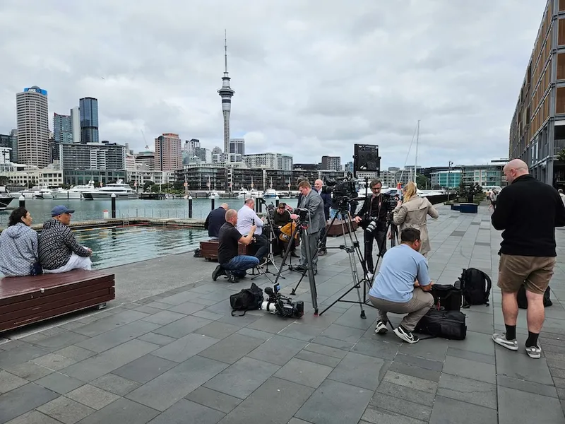 A group of journalists with cameras and equipment gather on a waterfront promenade in a city with modern buildings and a tall tower in the background. Some people sit on benches nearby. The sky is overcast.