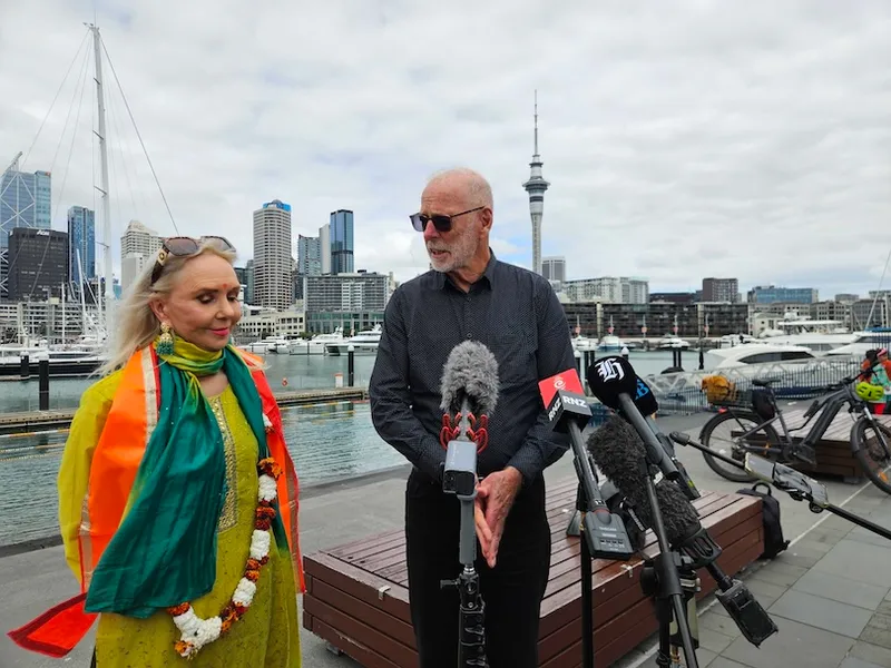 A woman and a man stand by microphones at a waterfront with boats and city buildings in the background. The man speaks while the woman listens, wearing a garland and sash. The Sky Tower is visible under a cloudy sky.