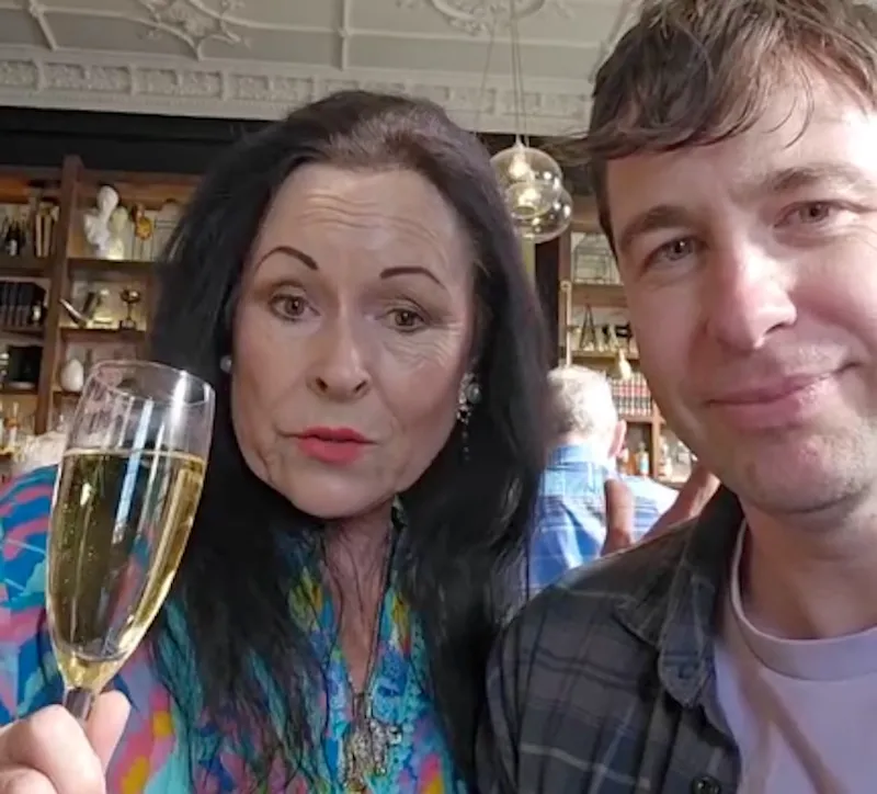 A woman with long dark hair holds a glass of champagne and poses next to a smiling man in a casual plaid shirt. They are indoors, with shelves and people visible in the background.