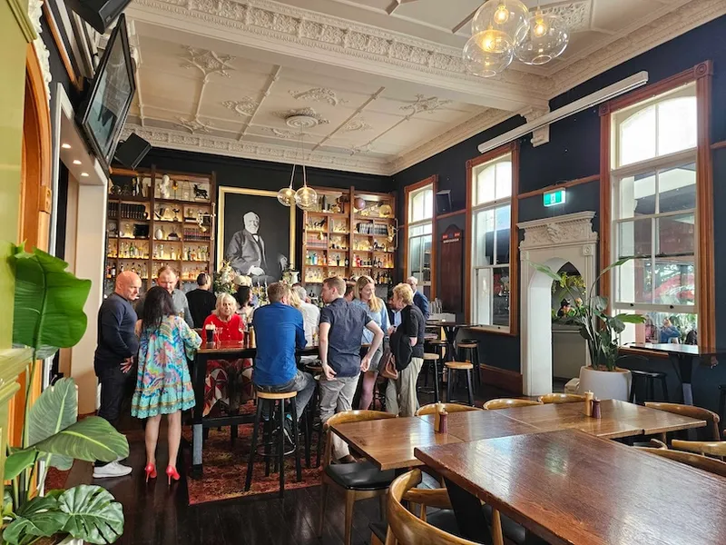 A group of people gather around a central table in a stylish bar with high ceilings, large windows, dark walls, and shelves full of bottles. A large portrait hangs on the back wall. Some tables and chairs are empty in the foreground.