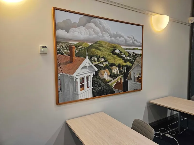 A framed painting of a hillside town with white houses and cloudy skies hangs on a light-colored wall above empty wooden desks and chairs in a modern, well-lit room.