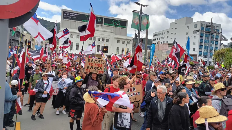 A large crowd of people is gathered in a street protest. Many are holding flags and signs. Several participants wear hats, and the background shows buildings and a partially cloudy sky.
