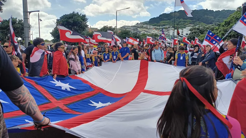 A group of people gathers outdoors, holding a large flag with red, white, and blue sections. Many are wearing blue and yellow sports jerseys, and others wave smaller red, white, and black flags. Trees and hills are visible in the background.