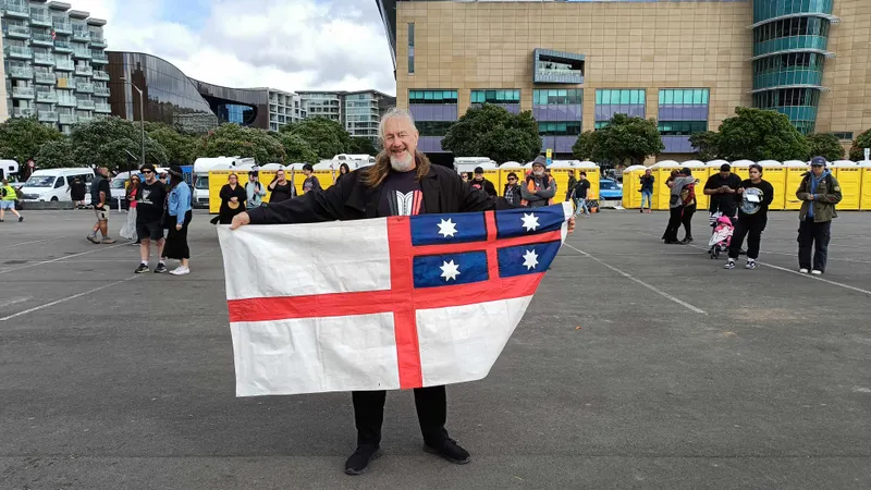 A person stands smiling in an open area holding a flag with a red cross on a white field and a blue canton with white stars. Several people are in the background, with a modern building and parked vans visible.