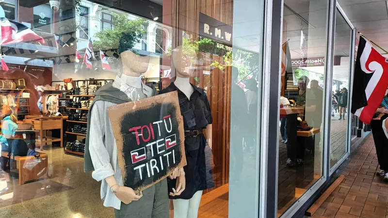 Two mannequins in a store display. One holds a sign with "TOITU TE TIRITI" in bold letters. The store is filled with clothing, and there's a New Zealand flag visible outside the window. Reflections of passersby are seen on the glass.