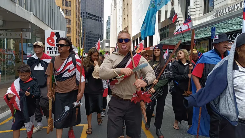 A group of people marches down a city street carrying flags and wooden staffs. One person in the centre sticks out their tongue. Buildings and signs are visible in the background. The group appears to be participating in a protest or demonstration.
