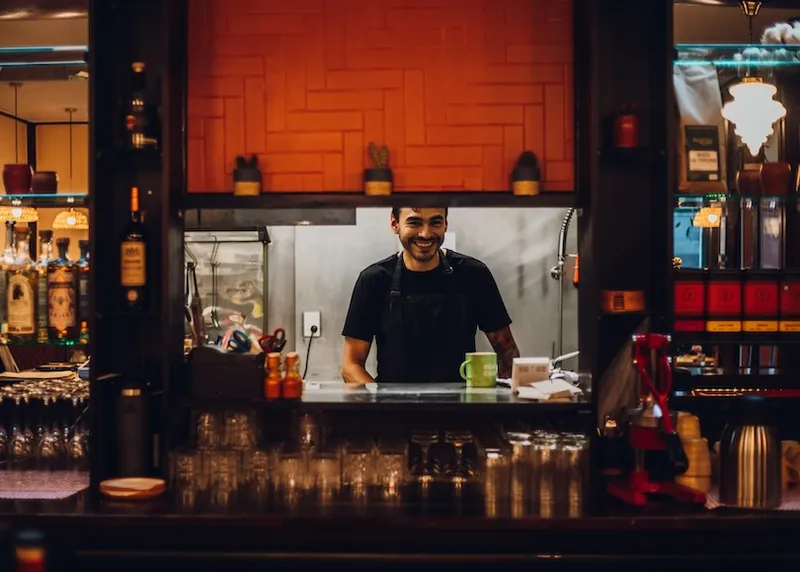 A smiling person in a black shirt and apron stands in a kitchen, viewed through a window from a bar area, with shelves of bottles, glasses, and small plants visible in the foreground.