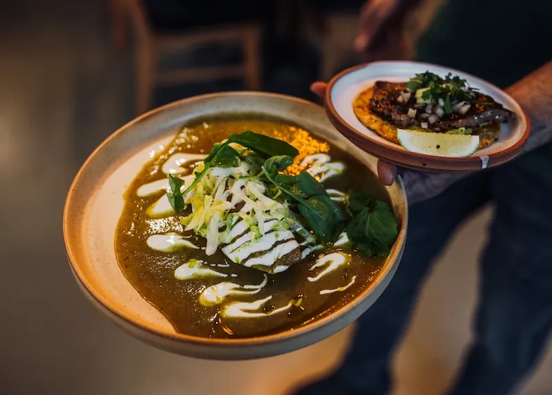A person holds a plate of a quesadilla frita covered in green sauce, cheese, and cream, garnished with greens, alongside a small plate with a taco, garnished with coriander and a lemon wedge.