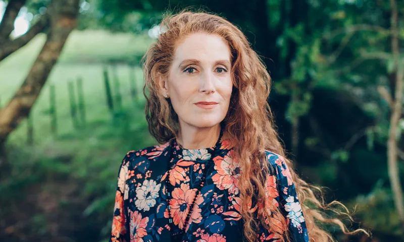 A photo of Catherine Chidgey who has long red hair and is outside in a shady area with trees and grass.