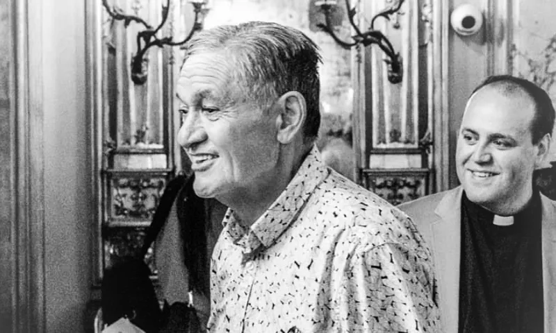 Two men smile and walk together in an ornately decorated room. The man in front wears a patterned shirt; the man behind him wears a clerical collar. The atmosphere appears joyful and warm.