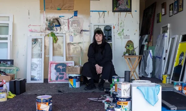 A person in black clothes sits in an art studio surrounded by paint cans, artworks, and art supplies, with colorful paintings and splattered walls in the background.
