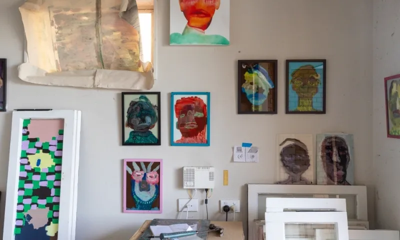 An art studio wall displays colourful, abstract portraits in frames. Below, a table holds papers and art supplies, with more framed artwork stacked nearby. A window at the top left lets in natural light.