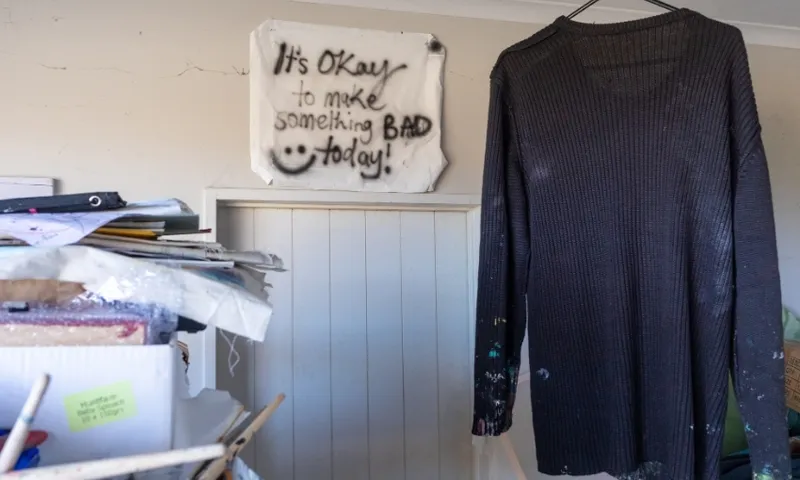 A long-sleeve dark shirt hangs near messy shelves with papers and art supplies. A sign on the wall reads, “It’s OKAY to make something BAD today!” with a smiley face.