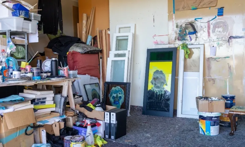 A cluttered artist’s studio with paintings of faces, scattered art supplies, canvases, paint cans, and frames leaning against the wall. Shelves and boxes are filled with various materials.