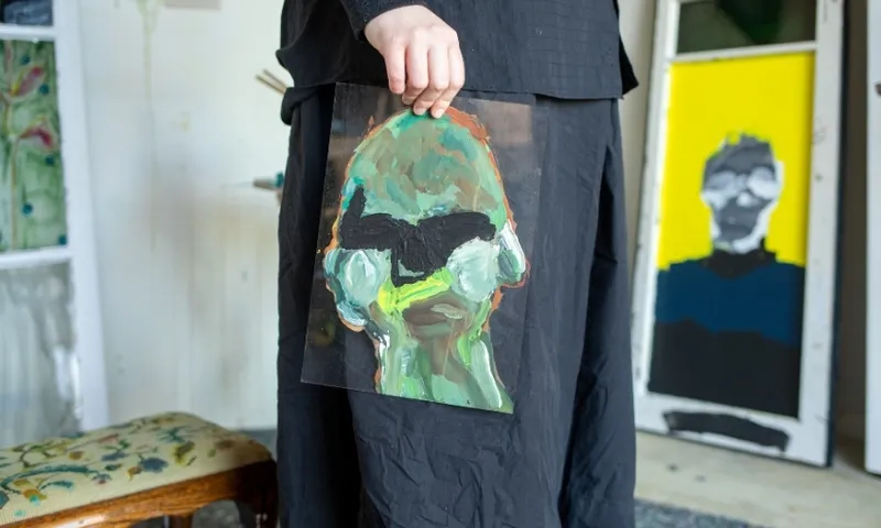 A person in black holds an abstract painting of a face with bold green, white, and black brushstrokes; another abstract portrait is propped in the background.