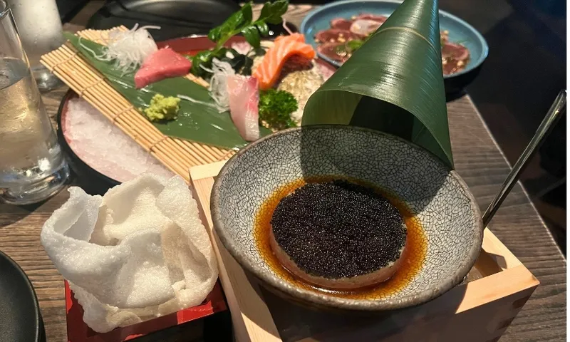 A beautifully plated Japanese meal with sashimi, assorted raw fish, wasabi, sauces, a bowl with caviar in soy sauce, rice crackers, and a decorative leaf on a wooden table.