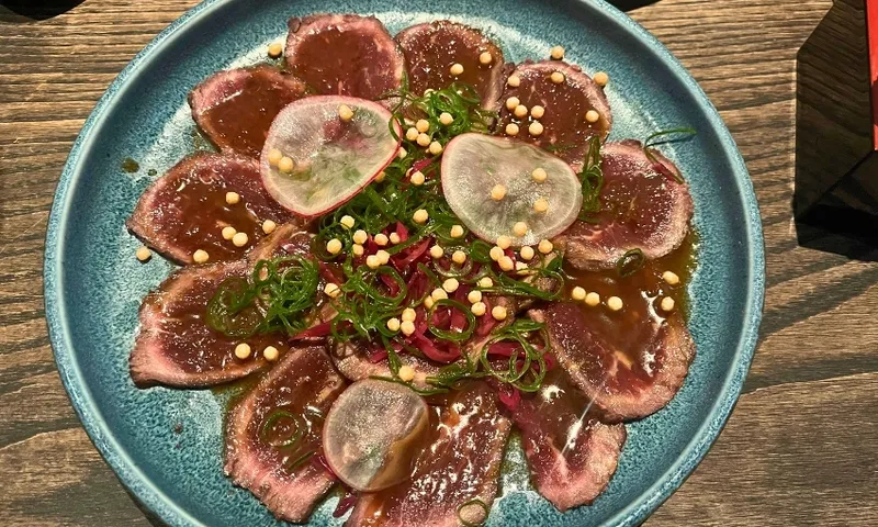 A blue plate with thinly sliced beef carpaccio, topped with sliced radish, green onions, crispy pearls, and a drizzle of sauce on a wooden table.