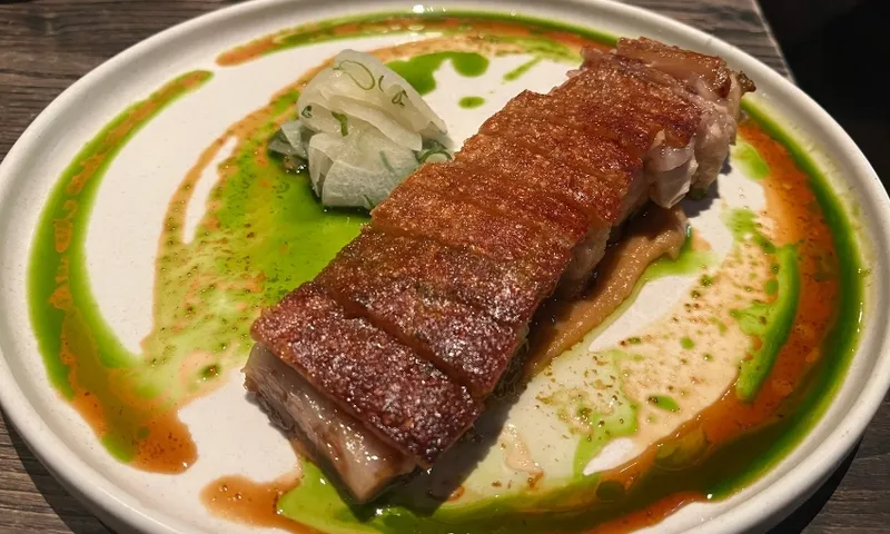 A rectangular piece of crispy pork belly sits on a plate with green and brown sauce swirls, accompanied by a small pile of thinly sliced pickled vegetables.