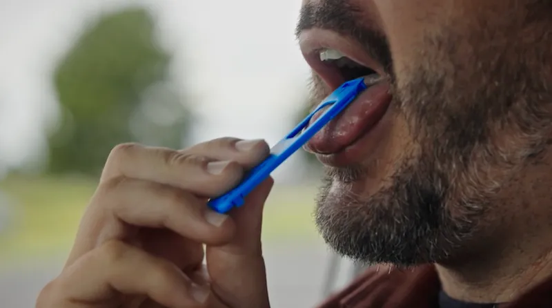 A close-up of a person holding a blue drug screening test to their tongue, with their mouth slightly open and facial hair visible.