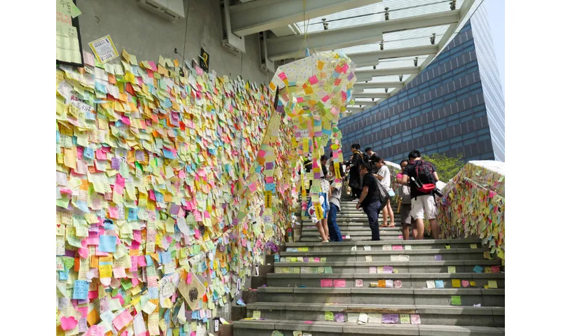 A photo of thousands of post it notes stuck onto a wall with stairs rising up beside it with people observing the notes.