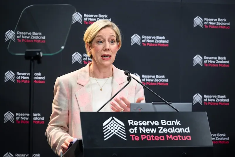 A woman speaks at a podium with “Reserve Bank of New Zealand Te Pūtea Matua” branding in the background. She gestures with one hand and appears to be addressing an audience.