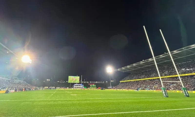 A brightly lit rugby league stadium at night, filled with spectators. The field is green with goalposts in the foreground and a large screen visible in the distance.