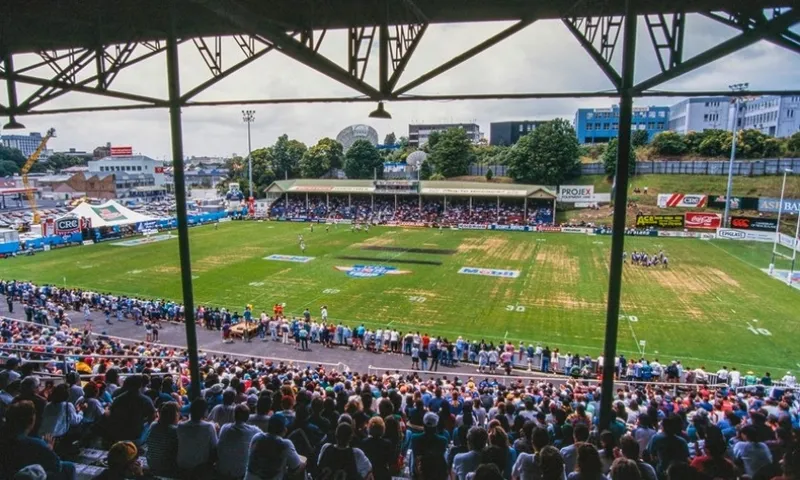 A wide view of a rugby league stadium with a green field, spectators in covered stands, and crowds along the sidelines. Buildings and trees are visible in the background under a partly cloudy sky.