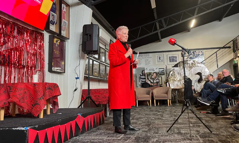 An elderly white woman wearing a long red coat holds a microphone to speak to a row of seated patrons.