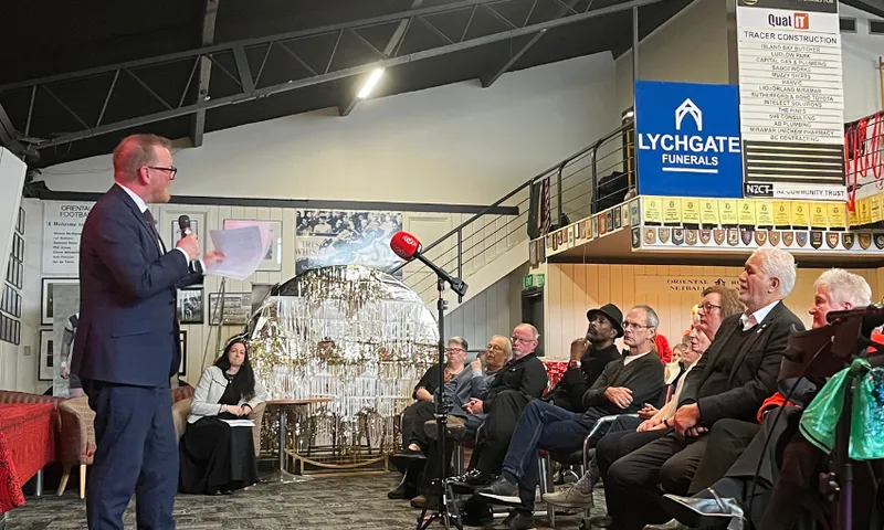 A middle-aged white man holds a pamphlet in one hand and a microphone to another as he speaks to a row of seated patrons.