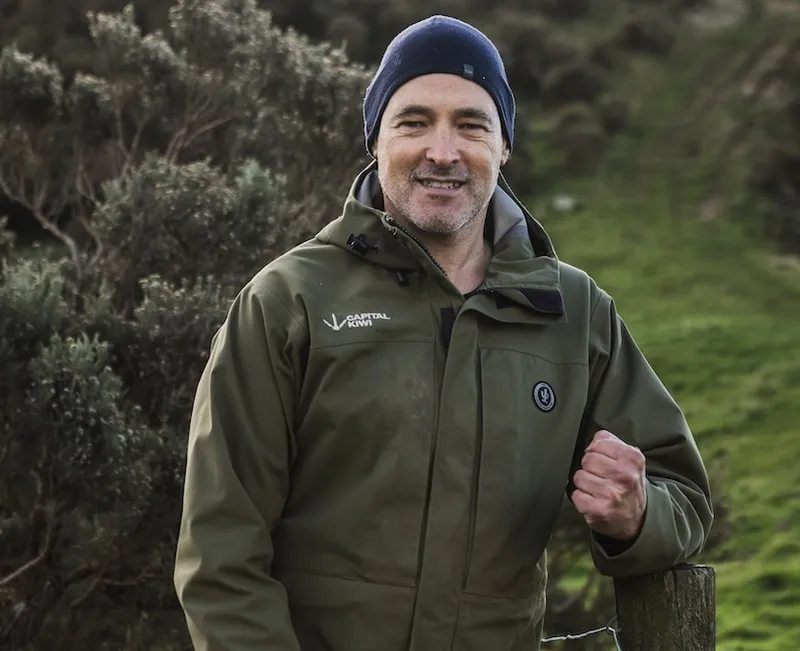 A man in a green jacket and navy beanie stands outdoors by a fence, smiling with greenery and shrubs in the background.