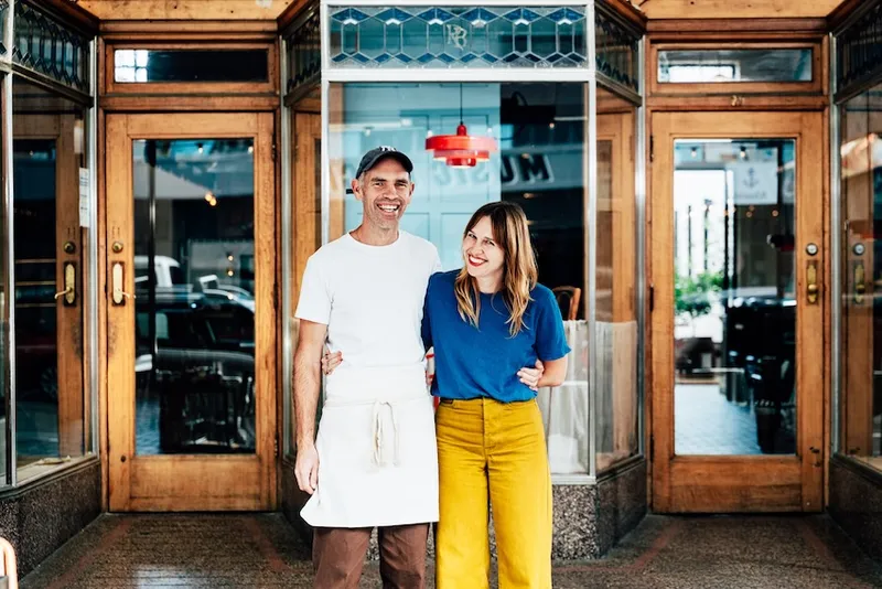 A man in an apron and a woman in yellow pants stand arm in arm, smiling, outside a restaurant with wooden doors and glass windows.