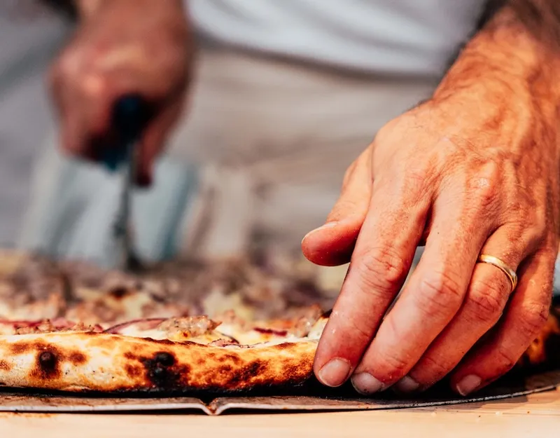 A close-up of a person’s hands, one holding a pizza and the other cutting it with a pizza cutter. The person is wearing a gold ring on their left hand. The pizza has a crispy crust and visible toppings.