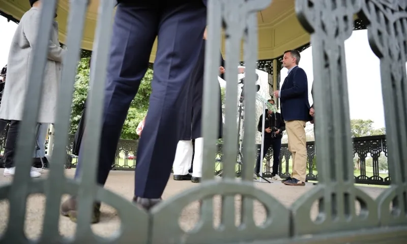 A group of people stand and talk inside a decorative gazebo, viewed from behind ornate metal bars. One person in a navy jacket and khaki pants faces others, with greenery visible in the background.