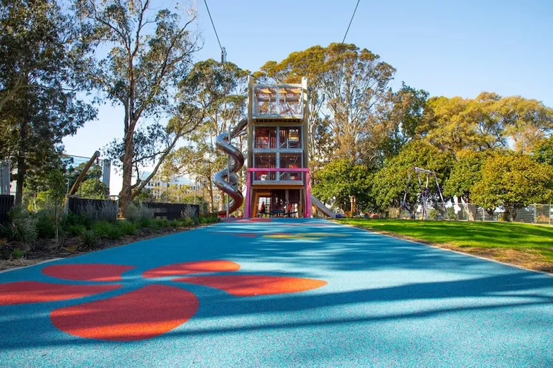 A tall playground tower with slides stands at the end of a blue play surface decorated with orange shapes, surrounded by trees and green grass under a clear sky.