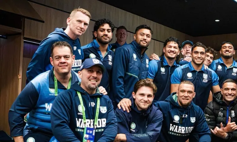 A group of men in matching blue New Zealand Warriors sportswear pose and smile together indoors, suggesting a sports team photo taken in a locker room or lounge area.