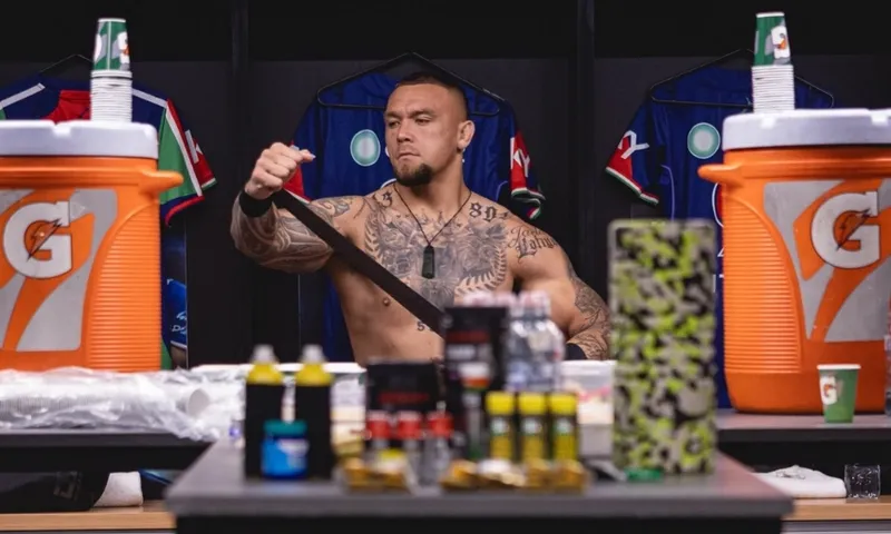 A shirtless, tattooed man sits in a locker room, wrapping his arm with tape. Sports drinks, water bottles, and equipment are on the table in front of him. Jerseys hang in the background.