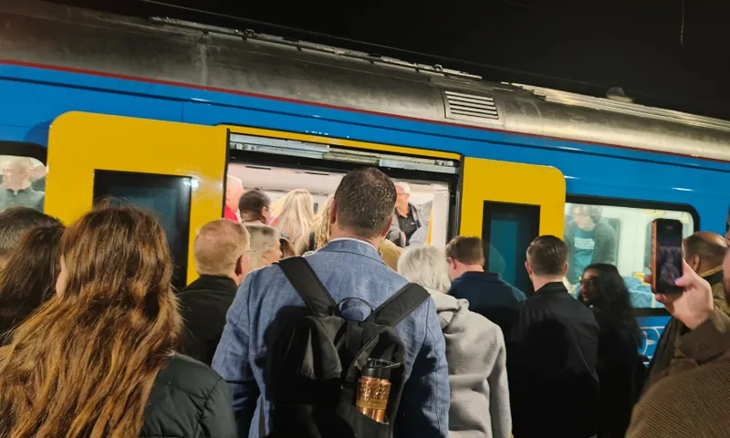 A crowd of people waits to board a blue and yellow train at night. The train doors are open, and passengers are visible inside. Some people are looking at their phones.