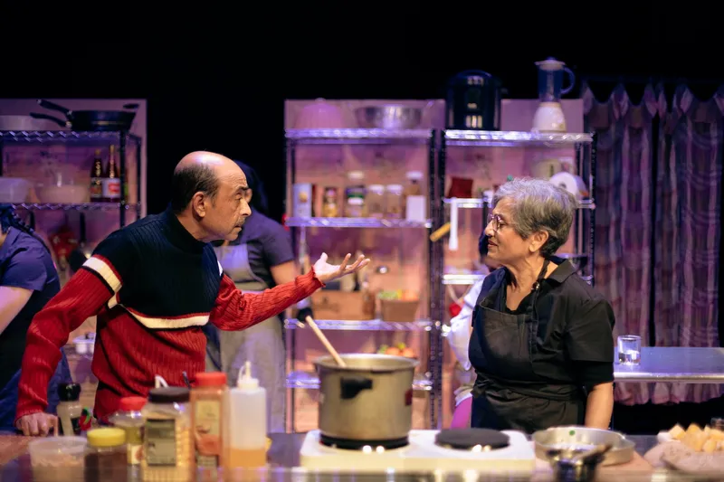 a stage play with a man and woman animatedly talking in a kitchen with a large pantry in the back and a large pot on the bench in front