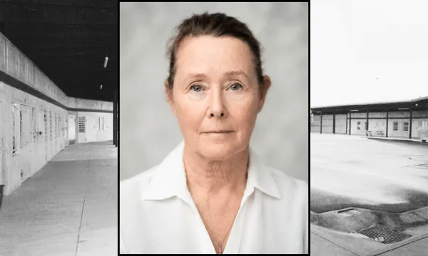 an older woman with white skin, blue eyes and brown hair pulled back in a bun stares sombrely at the camera. In the background a black and white photo of a prison yard