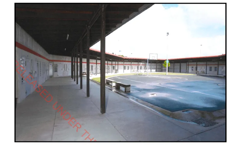 a photo taken on an old digital camera shows an open-air courtyard with a basketball hoop, concrete walkways and a rounded single story building surrounding it with cell doors