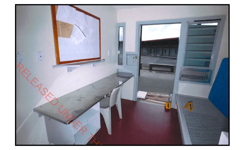 A digital camera photo taken with flash, showing the inside table, bed and open doorway of a prison cell with an open air courtyard outside