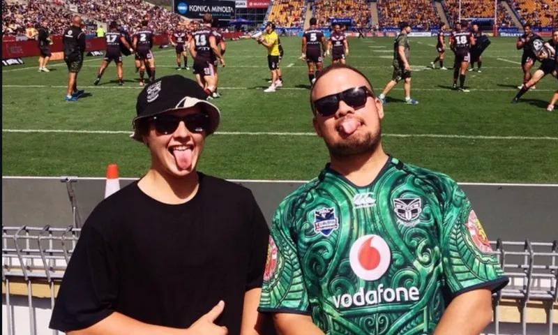 Two people stand in front of a rugby field, both wearing sunglasses and sticking out their tongues. The person on the right wears a green Vodafone Warriors jersey; rugby players and a stadium crowd are in the background.