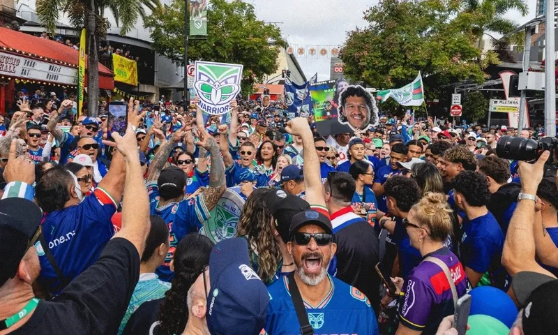A large crowd of people wearing blue jerseys gathers outdoors, cheering and holding up signs, including a Warriors logo and a large face cutout. Palm trees and buildings line the street in the background.