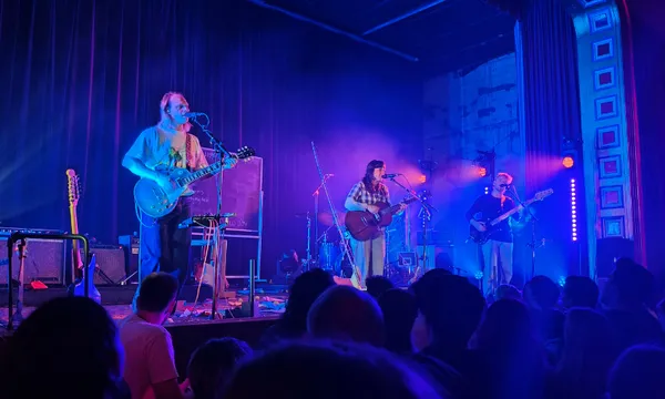 A band performs on a dimly lit stage with blue and purple lights, playing guitars and singing. The audience watches from below, and musical equipment is visible around the musicians.