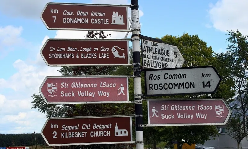 A signpost with directions to Donamon Castle, Stonehams & Blacks Lake, Suck Valley Way, Kilbegnet Church, Roscommon, and Athleague/Ballygar. Some signs are in both English and Irish. Trees in background.