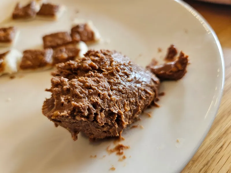 A close-up of a plate with a scoop of chocolate spread in the foreground and pieces of chocolate on white bread in the background.